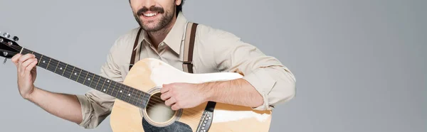 Vista recortada de hombre alegre con bigote en ropa de estilo retro tocando la guitarra acústica aislado en gris, bandera - foto de stock