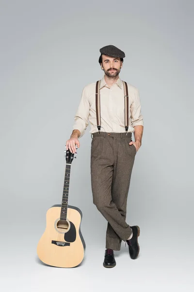 Comprimento total do homem com bigode em roupas de estilo retro e chapéu de pé com a mão no bolso perto da guitarra acústica em cinza — Fotografia de Stock
