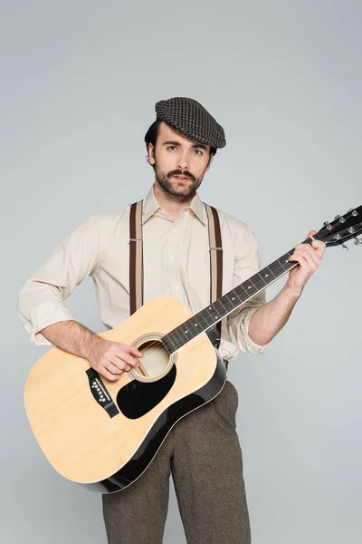 Человек с усами в ретро-одежде и шляпе, играющий на акустической гитаре, изолированной на сером — стоковое фото