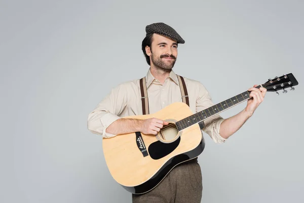 Hombre sonriente con bigote en ropa de estilo retro y sombrero tocando guitarra acústica aislado en gris - foto de stock