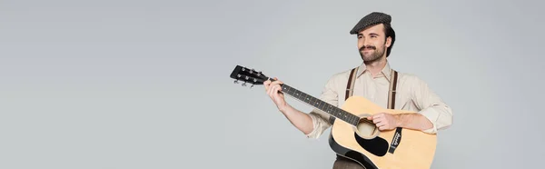 Усміхнений чоловік з вусами в одязі в ретро-стилі і капелюсі грає на акустичній гітарі ізольовано на сірому, банер — стокове фото