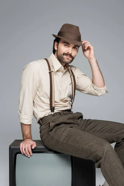 Hombre alegre ropa de estilo retro ajustando sombrero y sentado en la televisión antigua en gris - foto de stock