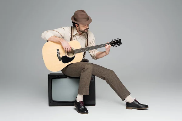 Pleine longueur de vêtements de style rétro homme jouant de la guitare acoustique et assis sur la télévision antique sur gris — Photo de stock