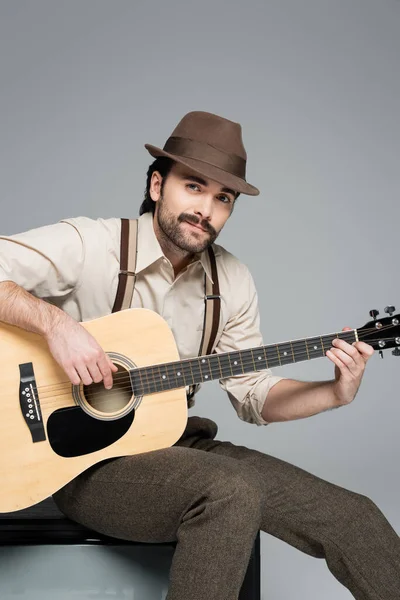 Hombre alegre ropa de estilo retro tocando la guitarra acústica y sentado en la televisión antigua en gris - foto de stock