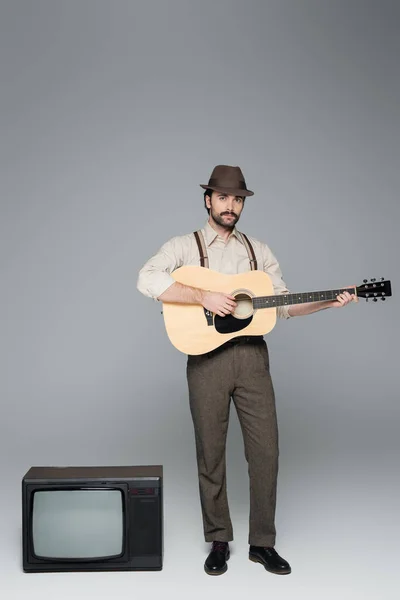 Full length of man retro style clothing playing acoustic guitar and standing near antique tv on grey — Stock Photo