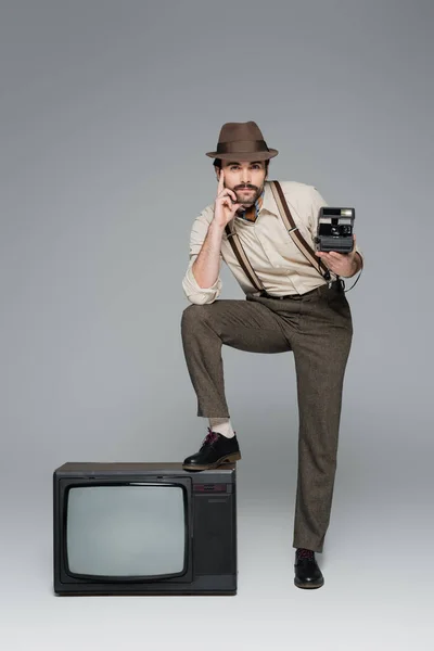 Pleine longueur de vêtements de style rétro homme et chapeau tenant caméra vintage près de la télévision antique sur gris — Photo de stock