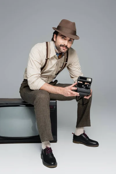 Piena lunghezza di uomo sorridente stile retrò abbigliamento e cappello con fotocamera vintage e seduto su tv antica sul grigio — Foto stock