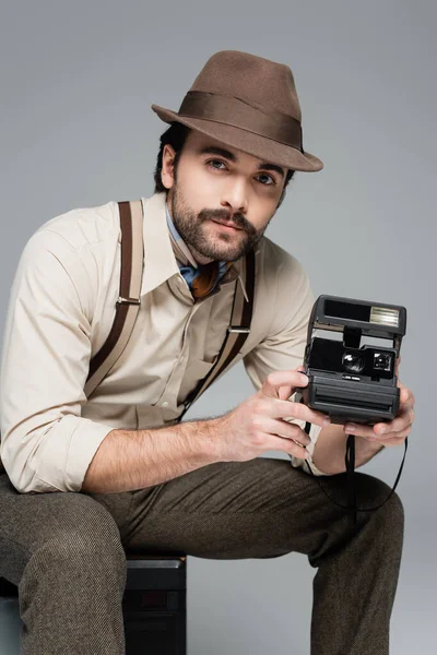 Man retro style clothing and hat holding vintage camera and sitting on antique tv isolated on grey — Stock Photo
