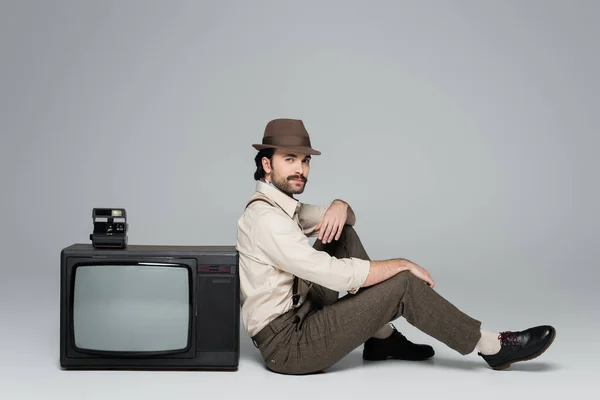 Одежда в стиле ретро в полный рост и шляпа, сидящая возле старинного телевизора с винтажной камерой на сером фоне — стоковое фото