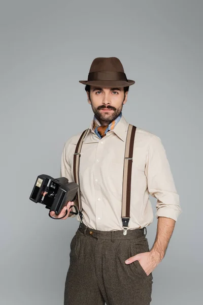 Человек в ретро стиле одежды и шляпы держа винтажную камеру и стоя с рукой в кармане изолированы на сером — стоковое фото