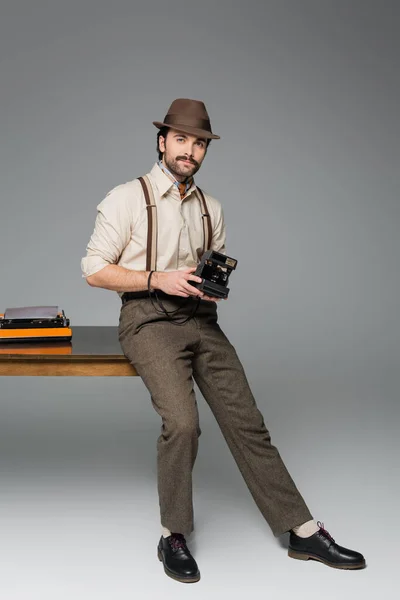 Pleine longueur de vêtements de style rétro homme et chapeau tenant caméra vintage et debout près du bureau avec machine à écrire sur gris — Photo de stock