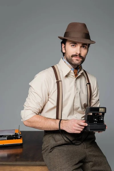 Man retro style clothing and hat holding vintage camera and standing near desk with typewriter isolated on grey — Stock Photo