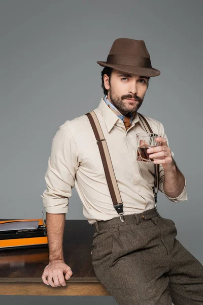 Hombre ropa de estilo retro y sombrero sosteniendo vaso de whisky y de pie cerca de escritorio con máquina de escribir aislado en gris - foto de stock