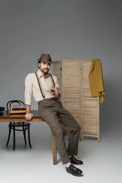 Мужская одежда в стиле ретро и шляпа держа стакан виски и стоя возле стола с пишущей машинкой на сером — стоковое фото