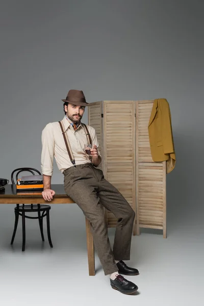 Man retro style clothing and hat holding glass of whiskey and standing near desk with typewriter on grey — Stock Photo