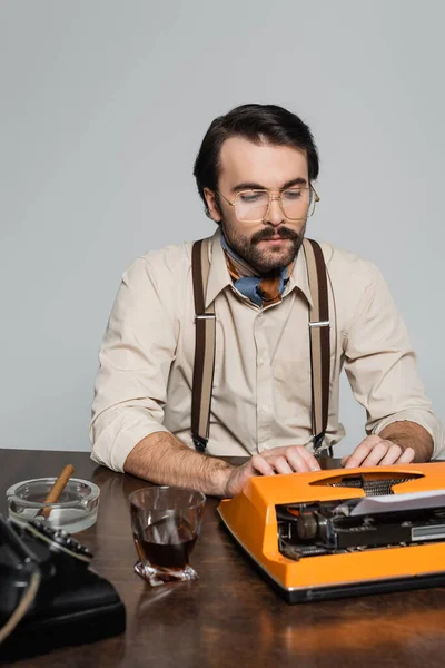 Journalist in eyeglasses using typewriter near glass of whiskey and cigar isolated on grey — Stock Photo