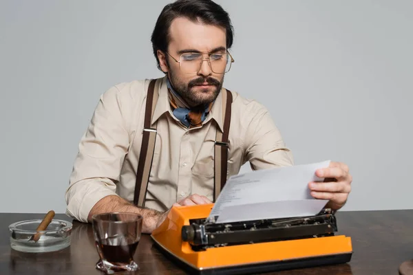 Journalist in eyeglasses looking at paper in typewriter near glass of whiskey and cigar isolated on grey — Stock Photo