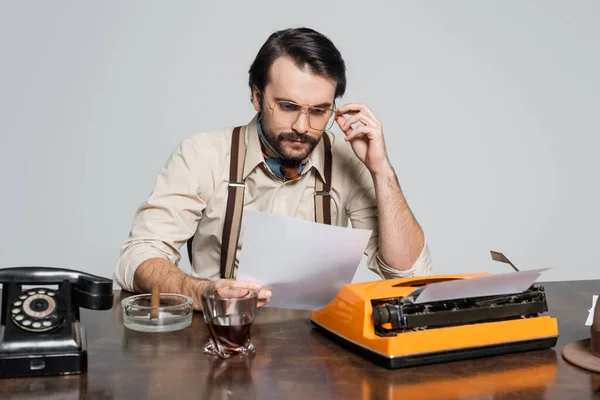 Journaliste à lunettes regardant du papier près d'une machine à écrire, verre de whisky et cigare isolé sur gris — Photo de stock