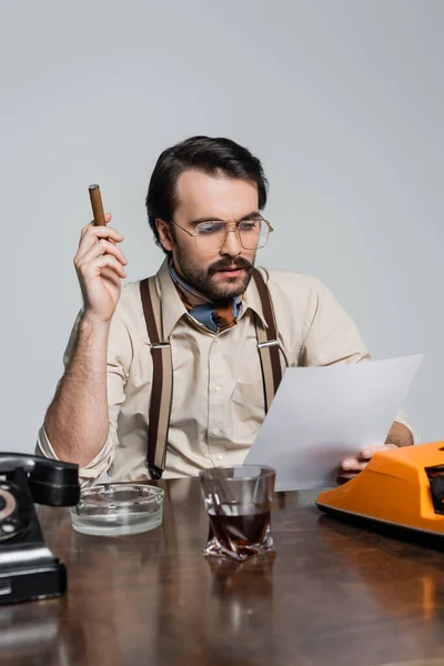 Journalist with mustache looking at paper and holding cigar near typewriter and glass of whiskey on desk isolated on grey — Stock Photo