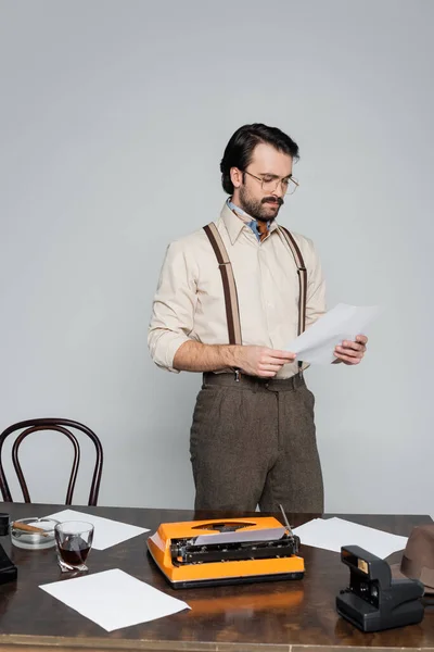 Journalist in eyeglasses looking at paper near typewriter, cigar, retro phone and glass of whiskey isolated on grey — Stock Photo