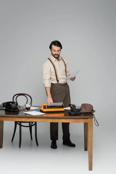 Journalist in eyeglasses holding paper near typewriter, glass of whiskey and vintage camera isolated on grey — Stock Photo