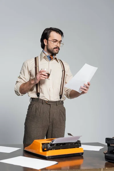 Periodista en gafas mirando papel y sosteniendo vaso de whisky cerca de la máquina de escribir en el escritorio aislado en gris - foto de stock
