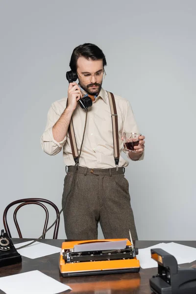 Jornalista em óculos segurando copo de uísque perto de máquina de escrever na mesa e falando no telefone retro isolado em cinza — Fotografia de Stock