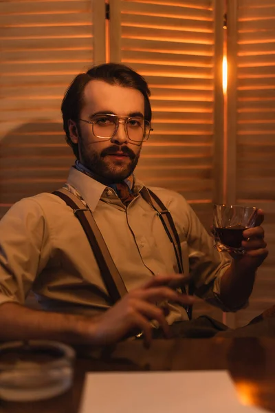 Hombre en gafas con vaso de whisky y cigarro cerca de pantalla plegable borrosa - foto de stock