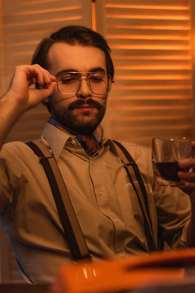 L'uomo regola gli occhiali e tiene il bicchiere di whisky vicino allo schermo pieghevole sfocato — Foto stock