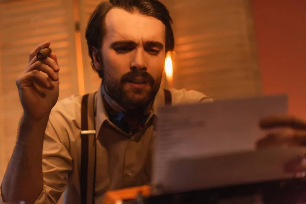 Writer holding cigar and looking at paper near typewriter machine against blurred folding screen — Stock Photo