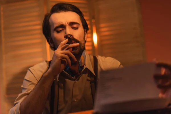 Writer smoking cigar and looking at paper near typewriter machine against blurred folding screen — Stock Photo