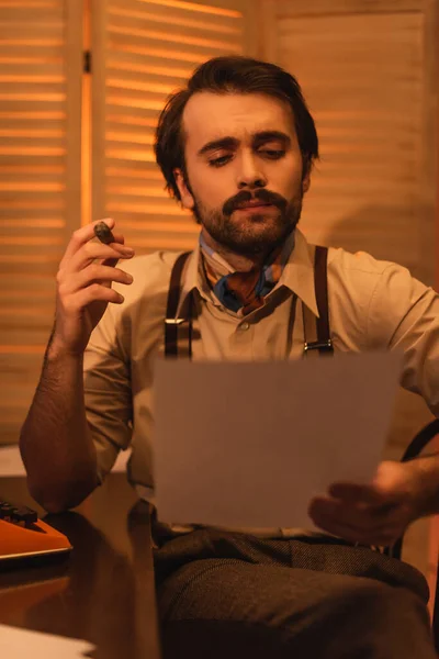 Homme avec moustache regardant le papier et tenant cigare près de machine à écrire rétro sur le bureau — Photo de stock