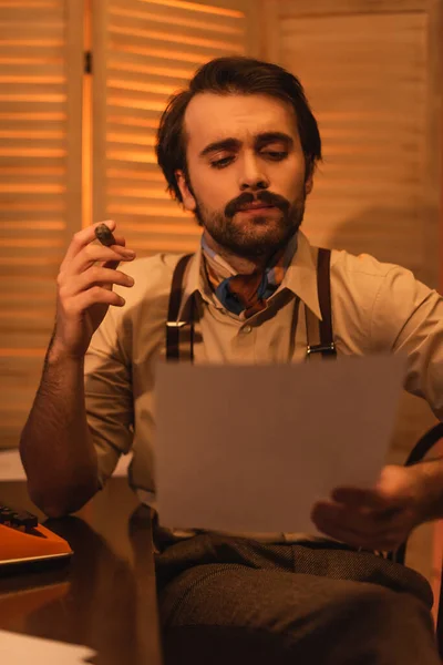 Man with mustache looking at paper and holding cigar near retro typewriter machine on desk — Stock Photo