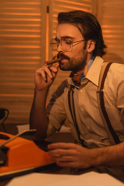 Writer with mustache and eyeglasses smoking cigar near retro typewriter machine on desk — Stock Photo