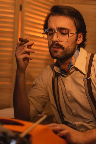 Jornalista com bigode e óculos fumando charuto perto de máquina de escrever retro borrada — Fotografia de Stock
