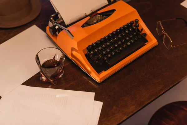 Vista de ángulo alto de la máquina de escribir naranja cerca de papeles y vaso de whisky - foto de stock