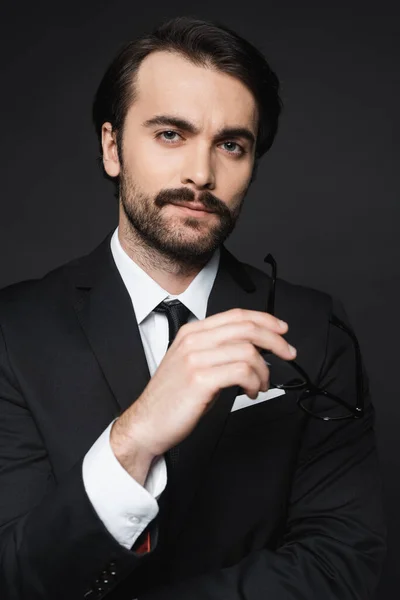 Businessman in suit holding glasses and looking at camera isolated on dark grey — Stock Photo