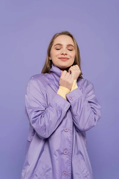 Pleased young woman with closed eyes in trench coat isolated on purple — Stock Photo