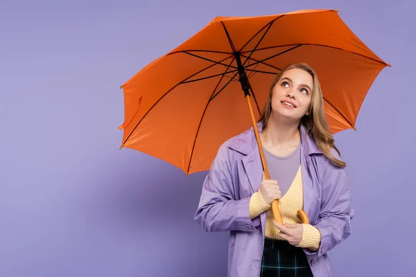 Mujer joven soñadora en gabardina de pie bajo paraguas naranja en púrpura - foto de stock