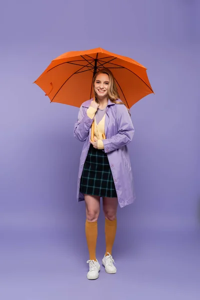 Full length of happy young woman in trench coat standing under orange umbrella on purple — Stock Photo