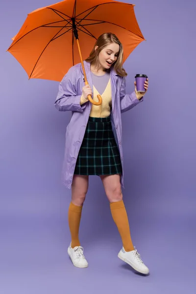 Pleine longueur de jeune femme étonnée en trench coat tenant tasse en papier et marchant sous parapluie orange sur violet — Photo de stock