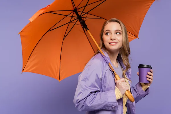 Jovem alegre em casaco de trincheira segurando copo de papel e de pé sob guarda-chuva laranja isolado em roxo — Fotografia de Stock