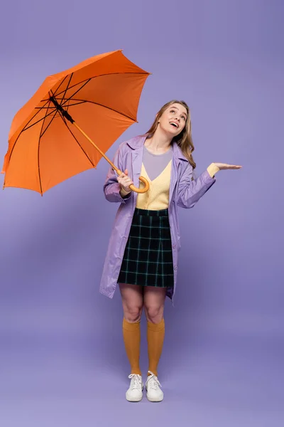 Volle Länge der fröhlichen jungen Frau im Trenchcoat gestikulierend, während sie einen orangefarbenen Regenschirm auf lila hält — Stockfoto