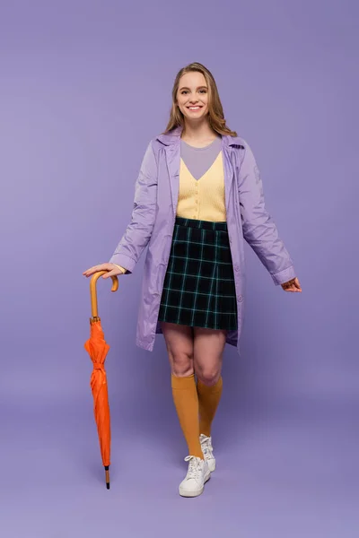 Full length of cheerful young woman in trench coat standing with orange umbrella on purple — Stock Photo