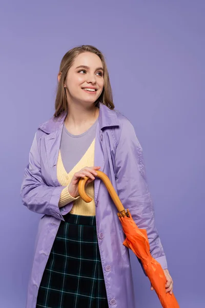 Allegra giovane donna in trench in piedi con ombrello arancione isolato su viola — Foto stock