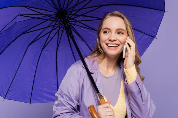 Happy young woman in trench coat standing with blue umbrella and talking on smartphone isolated on purple — Stock Photo