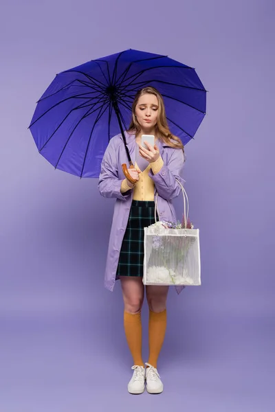 Full length of worried young woman in trench coat standing with blue umbrella and looking at smartphone on purple — Stock Photo
