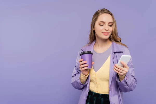 Jovem feliz em casaco de trincheira usando smartphone e segurando copo de papel isolado em roxo — Fotografia de Stock