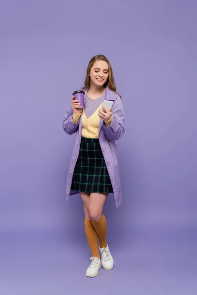 Full length of happy young woman in trench coat using smartphone and holding paper cup on purple — Stock Photo