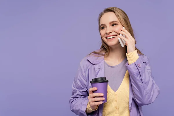 Sorrindo jovem mulher em casaco de trincheira falando no smartphone e segurando copo de papel isolado em roxo — Fotografia de Stock